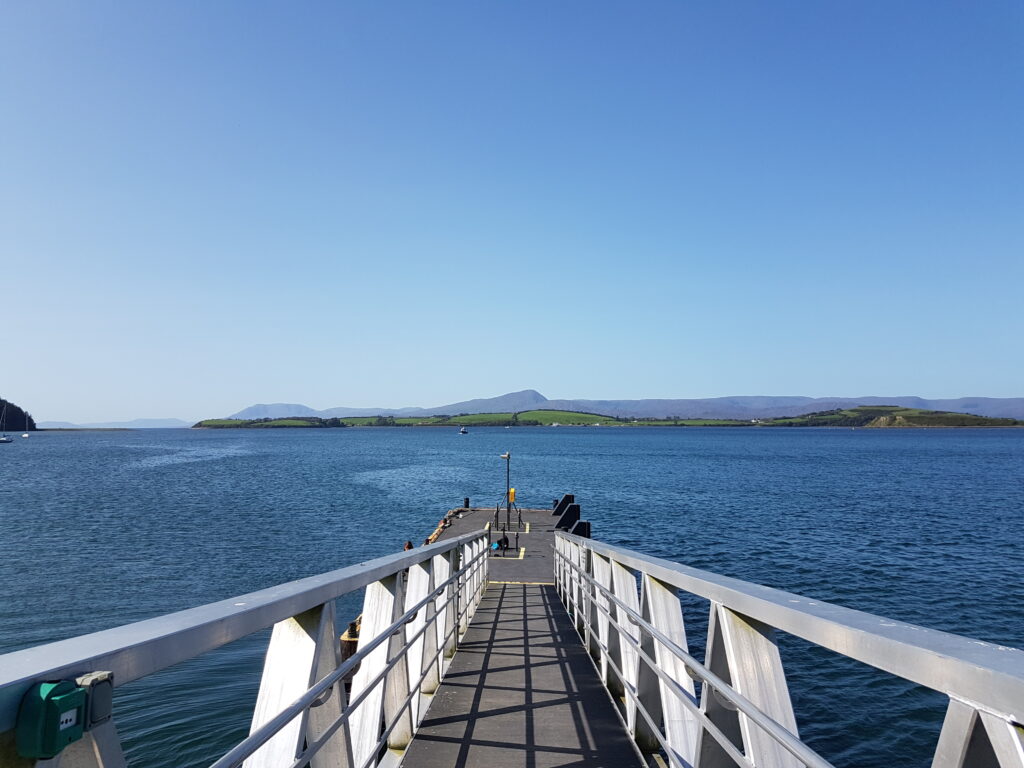 Colour photo taken from the top of a pontoon. The ramp leads down from the foreground to the pontoon, surrounded by calm dark blue sea. There is a low line of green hills on the horizon. The top half of the image is filled with blue sky.